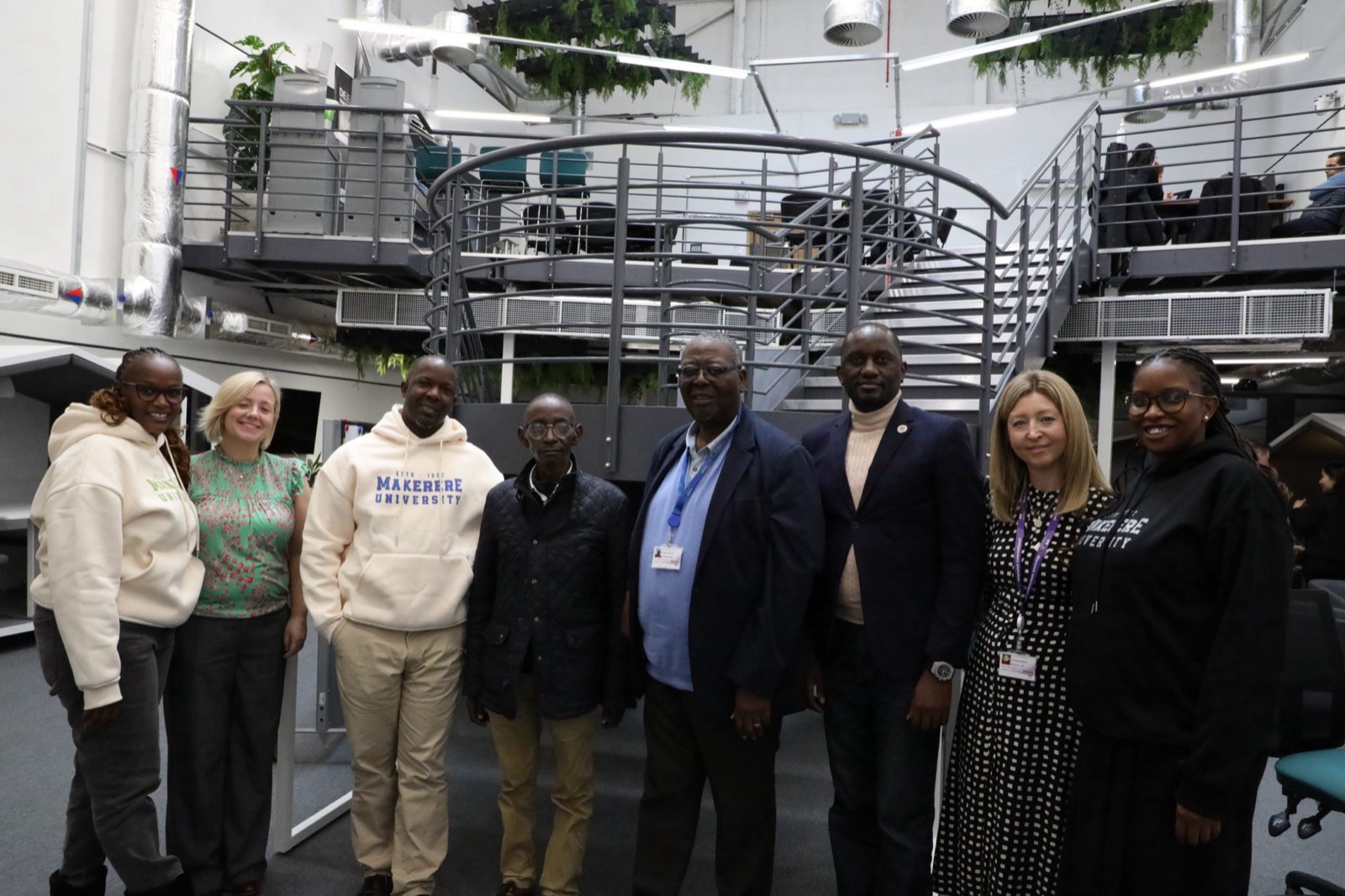 Nine people, several wearing Makerere University hoodies, standing together in a double-height workspace with a spiral staircase, mezzanine desks and hanging greenery.