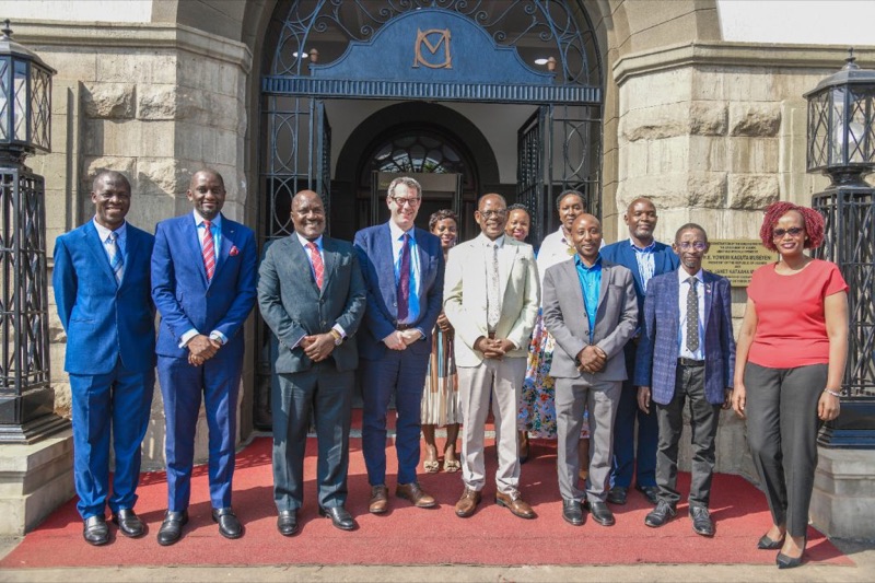 A group of about a dozen people in suits and formal dress standing in two rows on a red carpet at the arched stone entrance of a Makerere University building, beneath an ornamental wrought-iron gate.